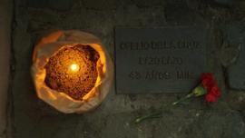 Vela encendida y clavel rojo junto a placa conmemorativa de Ofelio de la Cruz Lazo Lazo, DD. DD.